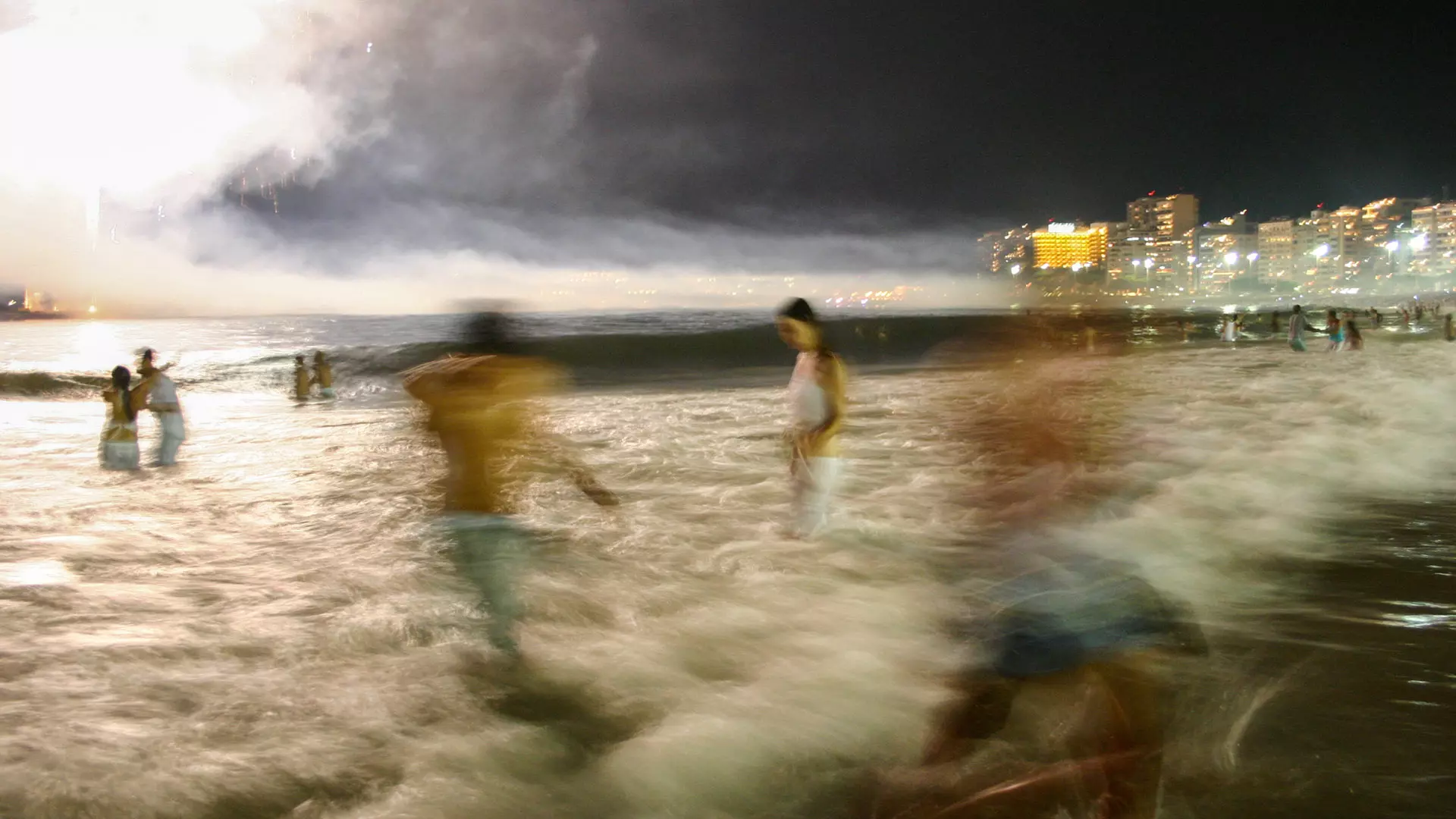 Pessoas vestidas de branco caminhando e pulando as ondas na praia à noite, com fogos ao fundo.